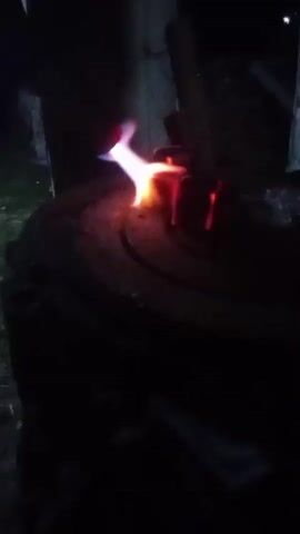 A dark, outdoor scene at night shows a close-up of a metal object being heated by a torch. The object, which appears to be made of dark metal and possibly has a rough surface, is glowing a bright orange and red in the intense heat. The torch, held by an unseen person, emits a bright blue and white flame that directly targets the object, causing it to melt or deform. The surrounding environment is dark and out of focus, with hints of indistinct shapes that could be structures or natural elements. The overall impression is one of intense heat and industrial or artisanal work in progress.
