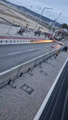 The video shows a white rally car speeding along a road course. The road has barriers on either side, separating it from a spectator area on one side and a sandy beach on the other. The car approaches from a "start" banner and a row of flames, then speeds away down the road. A person wearing a black jacket with BMW branding walks into the frame to watch the car. A safety marshal wearing an orange vest stands near a warning sign. The road is bordered by a cobblestone sidewalk with decorative patterns. The environment is urban, with buildings visible in the background under a slightly overcast sky. The camera position is elevated, likely from a building overlooking the racecourse.