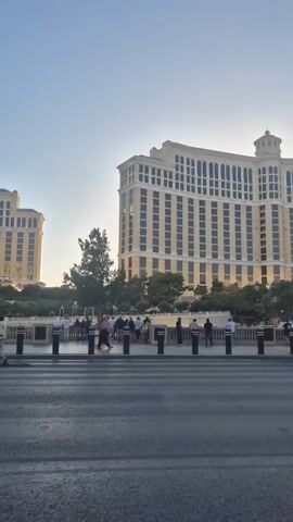 The video shows the Bellagio Hotel in Las Vegas, Nevada. The camera is stationary and captures the hotel's facade, fountains, and a group of people walking along the sidewalk. The sky is clear and light blue, suggesting it might be early morning or late afternoon. The street in the foreground is asphalt and appears wet. There are black bollards lining the edge of the sidewalk. The video provides a tourist-style snapshot of a popular Las Vegas landmark.