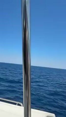 Here is a brief description of the video:

The video captures the serene view from a boat sailing on a vast, clear blue sea. The sky above is equally blue, creating a seamless horizon. A silver metal pole stands prominently in the foreground, acting as part of the boat's structure. As the camera pans slightly, other elements of the boat come into view, including a white surface and a collection of red and white ropes. The scene is set on a sunny day, highlighting the tranquility of the open sea and suggesting a peaceful journey.