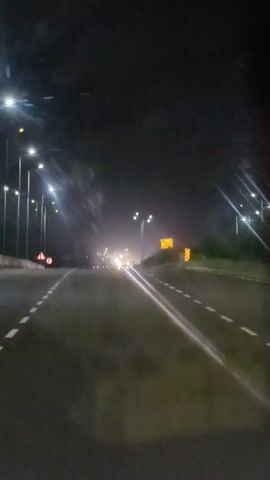 A first-person perspective video shows a nighttime drive on a highway. Streetlights illuminate the road and shoulder, where signs and barriers are visible. The road is wet, reflecting the lights, and a light fog or mist is present. In the distance, headlights and taillights of other vehicles are visible, suggesting traffic. The road has dashed white lines marking the lanes.
