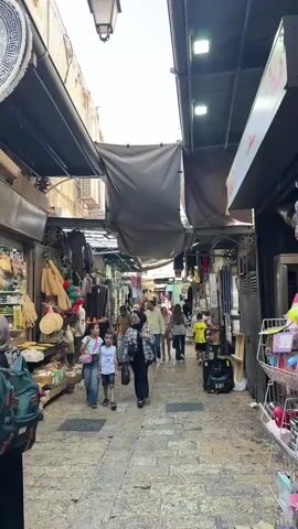 The video shows a bustling marketplace with people walking down a narrow cobblestone street lined with shops. In the foreground, a man wearing a white shirt and a black cap walks away from the camera. To his left, two young girls and a woman in a checkered jacket walk towards the camera. To the man's right, another man walks away from the camera carrying a bag of meat. Further down the street, several other people are visible, and shops display various goods. Towards the end of the video, the camera focuses on a vertical rotisserie with meat cooking, suggesting a food stall. The shops have awnings overhead, and the overall atmosphere is that of a lively, commercial area.