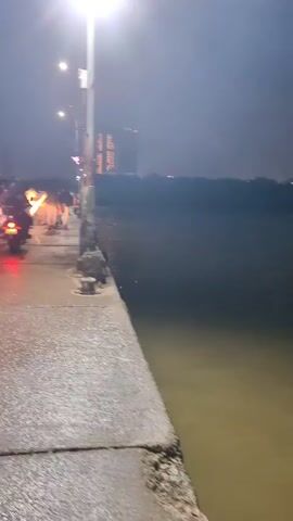 The video begins with a view of a wet, paved walkway at night, with a body of water to the right. A street light illuminates the area, casting reflections on the wet surface. A building with lights can be seen in the distance. Motorcyclists are present on the walkway, their headlights creating streaks of light. As the video progresses, a group of people, including children, are seen walking towards a boat docked at the pier. The boat appears to be a passenger ferry with a green and beige color scheme. Some people are boarding or disembarking the boat. The atmosphere is dark and appears to be overcast or rainy, given the wetness of the ground.
