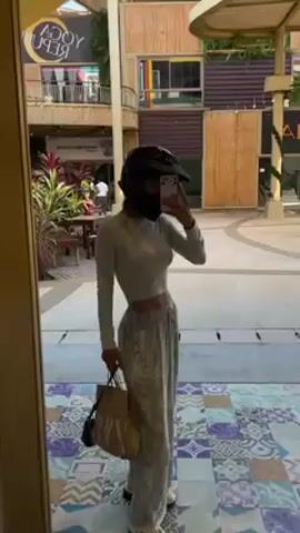 A woman in a motorcycle helmet takes a selfie in a mirror. The woman wears a long-sleeved crop top and patterned pants. She holds a brown tote bag. The mirror is situated in a public outdoor space featuring shops and restaurants. The floor is tiled with geometric patterns. Plants and outdoor seating are visible in the background.