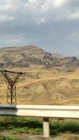The video shows a view from a vehicle moving along a road. In the foreground, a white guardrail with reflective blue and white stripes is visible. Beyond the guardrail, the landscape consists of dry, yellowish-brown hills and mountains with rocky formations and sparse vegetation. The sky is partly cloudy with a mix of gray and white clouds against a light blue background. The terrain appears arid and mountainous.