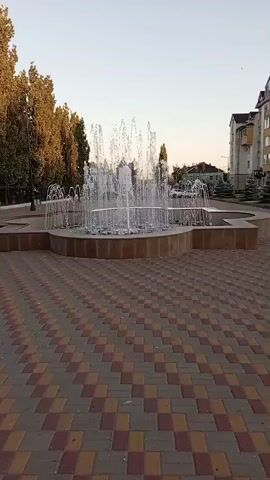 This video shows a public fountain in Kirsanov, Russia. The fountain is in the center of a paved area with a distinctive pattern of red and yellow tiles. Water sprays upwards in multiple streams, creating a dynamic display. Some of the jets have lights at their base, which appear to be changing colors, casting a subtle glow on the water.

In the background, tall trees with autumnal foliage line the left side of the frame, suggesting the time of year is likely fall. To the right, buildings characteristic of a town or city are visible, including a multi-story residential building with a light-colored facade. A few cars are parked or driving in the distance, indicating a typical urban scene. The sky is clear and bright, suggesting a pleasant day, and the soft light hints that it might be late afternoon or early evening.

There are no people directly visible in the foreground or actively interacting with the fountain in this clip. The focus is on the fountain itself and its surrounding environment. The overall mood is peaceful and serene, highlighting a moment of tranquility in a public space. No text is visible in the video.