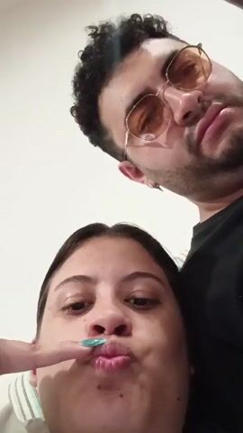 A low-angle, close-up selfie-style video shows two people, a man and a woman, posing for the camera. The man is in the background, wearing round, orange-tinted sunglasses and a black shirt. He has curly dark hair and is making a pouty face. The woman is in the foreground, also making a pouty face. Her right hand is raised, with her index and middle fingers extended in a peace sign. Her fingernails are painted a bright blue. The background appears to be an indoor setting with a plain, light-colored wall.