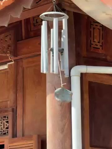The video shows a wind chime hanging outdoors, slightly swaying. The chime has a silver metal frame with multiple white cylindrical tubes. A stone or clay object hangs below, striking the tubes. The chime is affixed to a wooden building with intricate carved details. A white PVC pipe is visible near the chime. Sunlight illuminates the scene. The camera remains static throughout the short video.