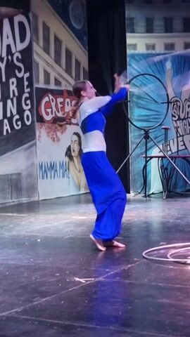 A woman with braided hair wearing a blue and white costume performs with a hula hoop on a stage. She is holding the hula hoop with her hands and spinning it around her body. The stage floor is dark, and the backdrop features painted murals of buildings and a bicycle. The lighting is dim, with spotlights illuminating the performer.