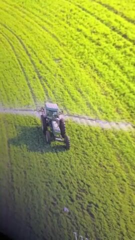 An aerial drone shot shows a green tractor spraying a field of crops. The tractor is moving across the field from left to right, leaving a trail of spray behind it. The field is very green and appears to be a field of young crops. The sun is shining, and the lighting is bright. The drone shot is slightly blurry due to the movement of the tractor and the drone.