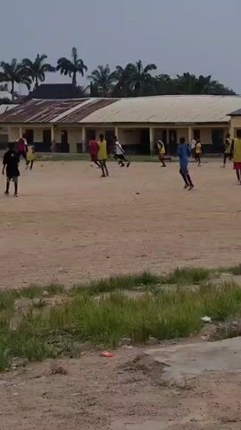 The video shows an outdoor scene of a soccer game. Several young people are playing soccer on a dirt field. In the background, there are buildings that appear to be schools or houses with corrugated metal roofs. Palm trees are visible in the distance. The sky is overcast. The players are wearing various colored shirts and shorts. The ball is visible on the ground at times. The camera is positioned at a distance from the game, offering a wide view of the field and surrounding environment. The game appears to be casual, with players scattered across the field.