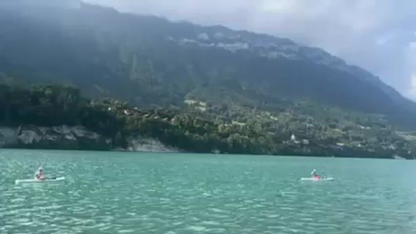 The video depicts a serene lake landscape, with two people on paddleboards in the foreground. The lake is a clear, turquoise color with gentle ripples on the surface. The people are paddling, moving slowly across the water.  Behind the lake, a lush, green mountainside rises steeply, with some buildings visible nestled in the trees. The sky is overcast, with a mix of clouds and sunlight.