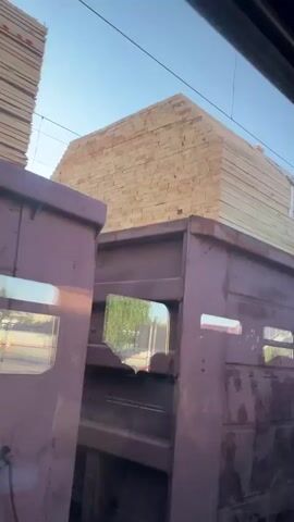 The camera is in motion, seemingly within a moving train. Through the train window, we see a flatbed railway car loaded with stacks of lumber. The lumber is secured with white straps and wooden supports. The railway car is painted in a faded purple color and has multiple window-like openings, but these appear to be structural features rather than actual windows. Text is visible on the side of the railway car, seemingly indicating ownership or registration information. The background features additional stacks of lumber and a clear, blue sky. Power lines are visible above.