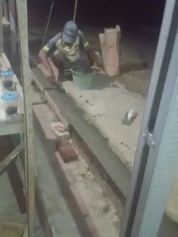 A person is working on a construction site. They are wearing a blue hard hat, a yellow and blue shirt, and a face mask. They are kneeling on the ground and using a trowel to apply cement to a structure. There is a bucket and a bag of cement next to them. The construction site appears to be outdoors and is lit by artificial light. The image is taken from an elevated perspective.