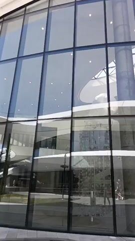 The video shows a walkthrough of the interior and exterior of a modern building with a curved glass facade. The interior features a large, open space with a central cylindrical column and spiraling staircases visible through the glass. Natural light streams in from the ceiling, illuminating the space. A few people are seen inside, walking or sitting on benches.

The exterior of the building is equally contemporary, with sleek, curved architecture and extensive glass panels reflecting the sky and surrounding landscape. The walkway outside is paved with large, grey rectangular tiles. Beyond the building, trees and rolling green hills are visible under a clear blue sky with scattered white clouds, suggesting a pleasant, sunny day. The overall impression is one of modern design and openness, with the building seamlessly integrating with its natural surroundings. The location is identified as Beregovoe, Russia.