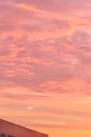 The video shows a sky filled with pink and orange clouds, likely at sunrise or sunset. The lower portion of the frame shows silhouettes of buildings and a street lamp with its lights on. There are no people visible in the video.