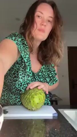 A woman with shoulder-length brown hair, wearing a green animal-print blouse, stands in front of a kitchen counter and displays a soursop, a green fruit with a spiky exterior. She cuts it in half and holds the two halves up to the camera, revealing the white flesh with black seeds. The background features a granite countertop, a white cutting board, and a stainless steel sink. There is a doorway visible behind her.