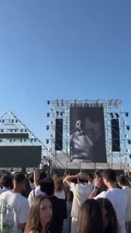 A video shows a large crowd of people watching a concert. The stage is large and elaborate, with a large screen showing a black-and-white image of a musician. The crowd is dense and excited, with many people raising their hands in the air. The sky is a clear blue.