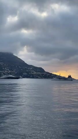 Two yachts are visible on calm water near a coastline with a mountain in the background. The sky is overcast with dramatic clouds, and a sliver of orange light suggests either sunrise or sunset. The yacht on the left is dark and sleek, while the yacht on the right is lighter in color and larger. Buildings are visible on the hillside. The water has subtle ripples.