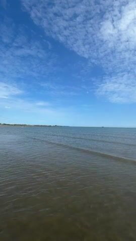 A low-angle shot shows a wide expanse of water under a bright blue sky filled with scattered white clouds. The water is a murky brown color and appears to be shallow, with small waves gently lapping at the shore. In the distance, a thin line of land is visible, with a few indistinct structures or trees. The overall scene is serene and natural, suggesting a peaceful day at the beach or by a lake. There are no people or specific objects in the frame, emphasizing the vastness of the natural environment.