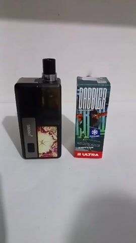The image displays two items on a light, plain surface, likely a table, against a light background. On the left is a dark-colored vaping device, possibly black or dark grey, featuring a transparent or translucent pod/tank on top. A decorative panel with a floral or abstract design in red and white tones is visible on its side. The brand name "SMOANT" is printed vertically on the device's side.

On the right, a rectangular box, likely containing e-liquid or vape juice, stands upright. The box is primarily dark grey or black with green and red accents. Prominent text on the box includes "DABBLER" in large stylized letters, "ХАЙПОВЫЙ" (Hyped), "ENERGETIK" (Energy drink), and "COOLER" accompanied by a snowflake icon. At the bottom of the box, "2 ULTRA" is visible in red.

At the 0:01 mark, a puff of white vapor is seen emanating from the top of the vaping device, suggesting it is being used or demonstrated. The scene appears to be indoors under artificial lighting.