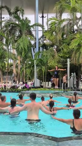 A group of people are participating in a water aerobics class in an indoor pool. A female instructor stands on a platform at the front of the pool, leading the class. The participants, dressed in swimwear, are arranged in rows in the pool, mirroring the instructor's movements. Palm trees and lush greenery surround the pool, creating a tropical ambiance. The ceiling of the facility is visible, with a modern, translucent design. The water is a vibrant blue, and sunlight streams in from above. The class appears to be energetic and engaging, with participants actively involved in the exercises.