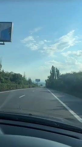 The video shows a view from inside a car traveling on a highway on a clear day. The road is paved and marked with white lines. Trees and greenery line both sides of the highway. A billboard is visible in the distance. The sky is blue with some scattered clouds. The dashboard of the car is partially visible in the foreground.