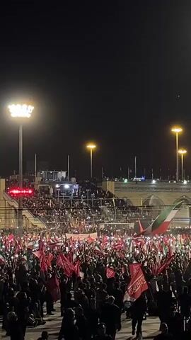 A nighttime scene shows a large crowd of people gathered outdoors, many holding up red flags and some holding up their cell phones with their lights on, creating a sea of illumination. The crowd appears to be in a large, open area, possibly a plaza or stadium, with tiered seating visible in the background. Several tall light poles with bright lights are scattered throughout the scene, illuminating the area. In the middle-right of the frame, a large flag with red, white, and green horizontal stripes is visible, which appears to be the flag of Iran. The overall atmosphere suggests a gathering or demonstration.