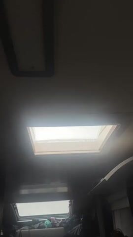 The video is a low-angle shot taken from inside a vehicle, possibly a van or RV, looking up through an open rectangular skylight. Outside, it is raining heavily, the skylight panels are translucent and the sky is an overcast white. The camera moves slightly, zooming in toward the skylight.