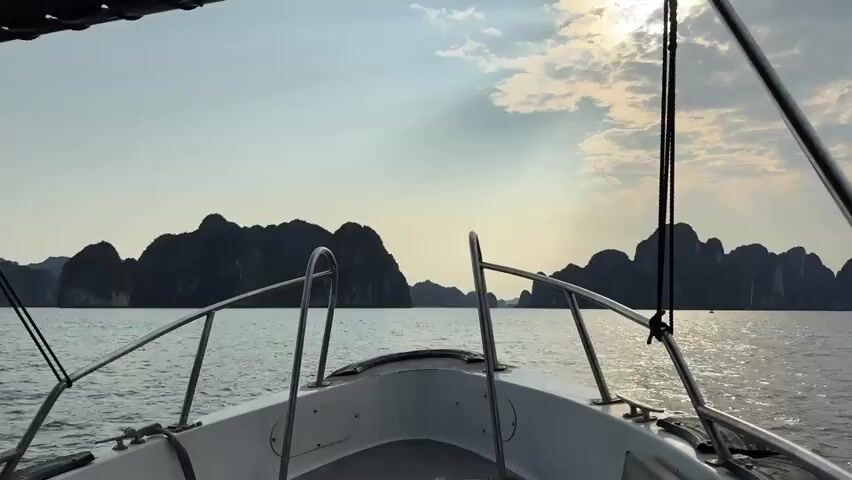 A boat is moving through water, likely a bay or sea. The view is from the front of the boat, looking out towards several islands. The islands are rocky and covered in vegetation. The sky is hazy with the sun partially obscured by clouds. The boat has a white hull and metal railings.