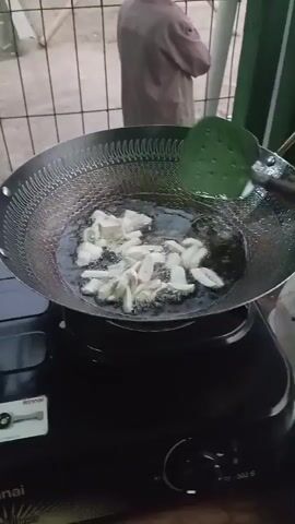 The video shows a person cooking, frying pieces of food in a wok. The wok is on top of a black stove. The food is white and cut into irregular shapes. The oil is bubbling and cooking the food. A green slotted spoon is being used to stir the food. There is a person in the background wearing a brown robe. The environment appears to be outdoors, possibly in a kitchen or cooking area.