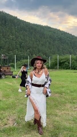 The video shows a woman in a cow-print jacket and a white dress, wearing a cowboy hat and jewelry. She is standing in a grassy field with a hill covered in trees in the background. There are other people and some equipment in the background, suggesting an outdoor event or gathering. The woman is looking at the camera and smiling, and at times she turns her head to the side. The sky is cloudy.