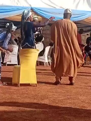 A large group of people stand under a white tent with blue and brown fabric draped around the edges. The ground beneath them is covered in reddish-brown dirt. In the foreground, a man in a brown traditional outfit with an embroidered collar faces a woman in a long, dark blue dress, a light blue patterned headwrap, and an embroidered top. Another woman in a dark patterned dress stands to the right of the man. To the left of the woman in blue, another woman in a light blue patterned dress bends over to pick something up from the ground next to a yellow chair. Other people can be seen in the background.