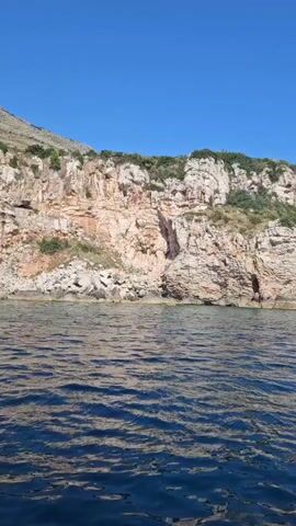 The video shows a boat traveling on the water, with a rocky cliff face to the left and a clear blue sky above. There are several people visible on the boat, with one person sitting on the edge, holding a drink. The boat is creating a wake as it moves through the water. The cliff face has some vegetation growing on it, and a modern white building is visible at the top of the cliff. The overall impression is a scenic view of a coastal area on a sunny day.