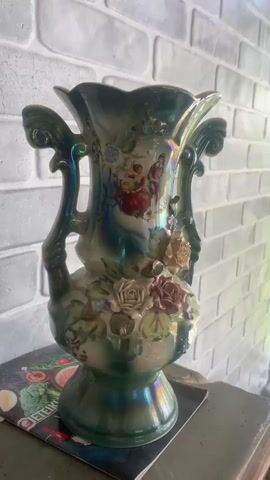 The video pans across a decorative vase that is green with iridescent highlights. The vase has handles and is adorned with raised roses and painted scenes of people in historical attire. It is placed on a table or surface in front of a brick wall.