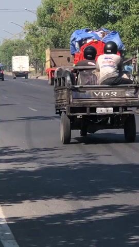 A traffic scene unfolds on a sunny day. A flatbed truck, possibly a Viar brand, moves down a paved road. Three individuals are seated in the back of the truck. One person, wearing a white helmet and a light-colored shirt with dark pants, faces away from the camera. Another person, also in a white helmet, wears a dark shirt. The third person is partially obscured. The truck bed contains a large red container covered with a blue tarp. In front of the truck, a person wearing a red shirt and a black helmet rides a motorcycle, also facing away from the camera. To the right of the truck, another person wearing a cream-colored shirt and a light-colored hat rides a scooter, facing away from the camera. Further ahead, more vehicles, including other motorcycles and trucks, are visible on the road. The environment appears to be a moderately populated area with trees lining the sides of the road and utility poles visible in the background. The sky is clear and blue. The road surface shows tire marks.