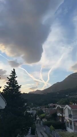 The video shows a view of a town and mountains under a cloudy sky. There are several contrails from airplanes forming a pattern in the sky. The town has small houses with tiled roofs. A tall evergreen tree is visible on the left side of the frame. Mountains covered in trees are in the background. The sky has a mix of light blue and dark gray clouds. Some indistinct voices can be heard in the background.