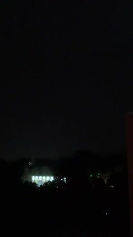 The video captures a night scene with dark skies and a distant building illuminated by lights. In the center of the frame, a bright yellow lightning bolt streaks across the sky, illuminating the dark clouds. The lightning is the most prominent event in the short clip.