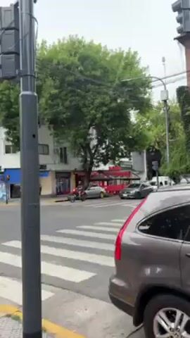 The video shows a street corner in a city, likely during the daytime. A red dirt bike is driven across a crosswalk. Several other vehicles are visible, including cars and a van. Pedestrians are walking on the sidewalks. Buildings and trees line the street, with a traffic light pole in the foreground. The sky appears overcast.