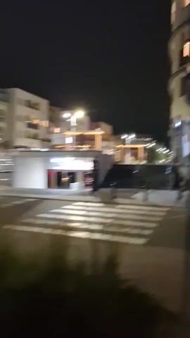 The video begins with a view of a pedestrian crossing at night. A building with lit windows is visible to the right. The camera then pans to the left, showing a street with cars parked and moving, and a multi-story building with glowing windows. The view shifts again to a sidewalk in front of a row of businesses, one of which has a green sign with the word "natura" on it. The video ends with a view of the sidewalk at night, with red lighting emanating from some of the businesses.