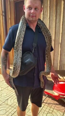 A young man stands outdoors, smiling and looking off-camera, with a large python draped around his neck and shoulders. He is wearing a dark blue short-sleeved collared shirt, dark shorts, and a black shoulder bag with "MARKBERG" visible on it. The python is mottled with brown and beige patterns, resting heavily on his upper body. The man appears relaxed and happy. The setting includes light-colored, patterned tiled ground and a wooden wall or building in the background. A red scooter is partially visible in the background on the right. The scene takes place in Thới Sơn, Vietnam.