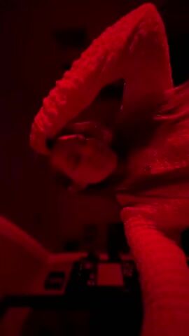 A video shot from a low angle shows a person lying down, illuminated by a red light. The person appears to be female, with long hair, and is wearing a textured long-sleeved top. She moves her arms slightly. In the background, dimly lit, are a wall, a small desk, and some objects on it. The red lighting dominates the scene, obscuring details.