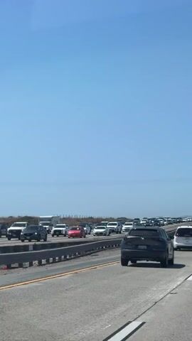 The video shows a traffic jam on a highway. The camera is in a vehicle moving slowly through the congestion. Numerous cars and trucks are visible, bumper-to-bumper, in multiple lanes. The sky is clear and blue. The environment appears to be a flat, open area with some vegetation or distant structures visible on the left side of the road. The time stamp indicates the video was recorded on a sunny day.