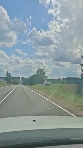 The video shows a view from a car driving on a paved road through a rural landscape on a sunny day with scattered clouds. The road has two lanes with white lines marking the edges and a double white line dividing the lanes. On the right side of the road, there is a green field, trees, and a road sign with some writing in a foreign language and a symbol of a village. On the left side of the road, there is a field with crops, and trees. Several vehicles are visible on the road ahead, including a white van and a red car. The sky is blue with large white clouds. The camera is positioned inside the car, looking forward through the windshield.