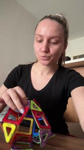 A woman is sitting at a wooden table, focusing on assembling a multi-colored, magnetic, geometric structure. She has light skin, her blonde hair is pulled back, and she wears a black V-neck shirt. The toy consists of colorful magnetic shapes, primarily triangles and squares, connected to form a sphere-like structure. The colors include red, yellow, blue, green, and purple. The woman's fingers are visible, adorned with light blue nail polish. The background includes a light-colored wall and a cabinet. Sounds of speech are heard in the background.