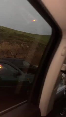 From the interior of a car, the video shows a rainy scene alongside a road where a group of individuals are gathered near parked cars. A grassy hill is visible in the background. Raindrops streak the car's window, obscuring the view slightly. The group of people appear to be in conversation near the vehicles. The sky is overcast and the lighting is dim. The focus is on the people and cars beside the road with a slight emphasis on the landscape behind them.