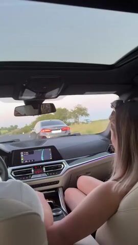 The video captures the interior of a modern car, likely a BMW, with a panoramic sunroof. The camera is positioned from the back seat, looking forward. In the foreground, the back of a person's head and shoulders are visible on the left, with their arm resting on the seat. On the right, a woman with blonde hair, wearing sunglasses on her head, is seated and looking towards the front of the car. Her legs are crossed and visible in the frame. The car's dashboard is prominent, featuring a large digital display showing navigation, and various buttons and controls with ambient lighting. The driver's side steering wheel and part of the digital instrument cluster are visible. In the mid-ground, through the windshield, another car, appearing to be a grey sedan, is driving ahead on a winding asphalt road. The exterior environment shows a landscape with greenery and trees, suggesting a rural or semi-rural setting. The sky appears overcast or dusk. The car is in motion, and the perspective shifts slightly as it navigates the curves of the road.