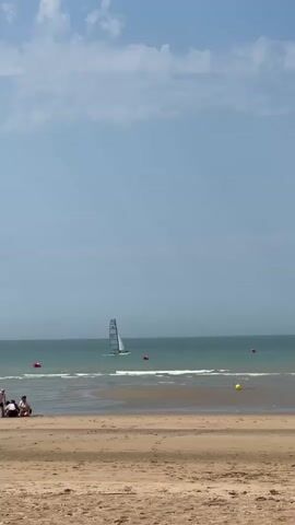 A wide shot shows a sandy beach with gentle waves lapping the shore under a partly cloudy sky. In the distance, a sailboat with a white sail navigates the green water. Several red and yellow buoys are visible in the water. On the beach, a small group of people are seated near the water's edge on the left side of the frame, while another person walks along the wet sand in the middle ground. The focus is on the tranquil beach scene with the sailboat as a distant element.