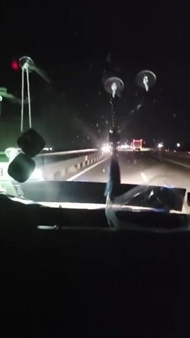 The video shows a view from inside a vehicle at night, looking through the windshield at a highway. The headlights of oncoming vehicles create streaks of light in the darkness. There are two fuzzy dice hanging from the rearview mirror on the left side of the windshield. On the dashboard, there is a clear plastic covering with some items underneath, possibly papers or a map. A dark, blue, tasseled ornament hangs from the center of the windshield. The highway is visible in the distance, with a barrier separating the lanes and several vehicles, including what appear to be trucks, driving on it. The overall impression is that of a night drive on a highway.