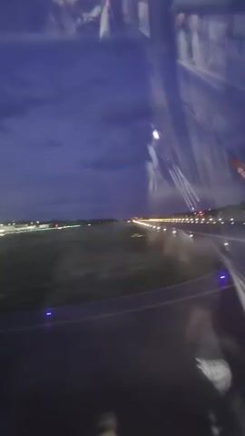 The video shows a view from inside an airplane as it is landing at night. The runway is visible, with rows of bright white and green lights. The wing of the airplane, with the EasyJet logo, is partially visible in the lower right corner of the frame. The sky is dark blue. Reflections of people inside the airplane are visible in the window.